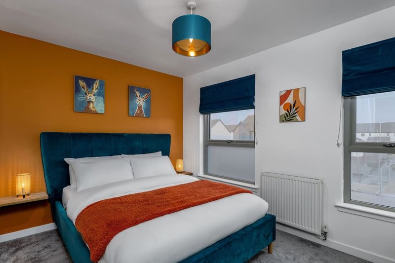 Modern bedroom with teal upholstered bed, orange accent wall, pendant light, and city views from windows