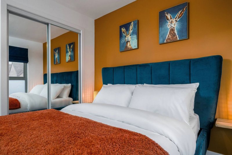 Modern bedroom with teal upholstered headboard, white pillows, orange throw, and framed artwork on mustard wall