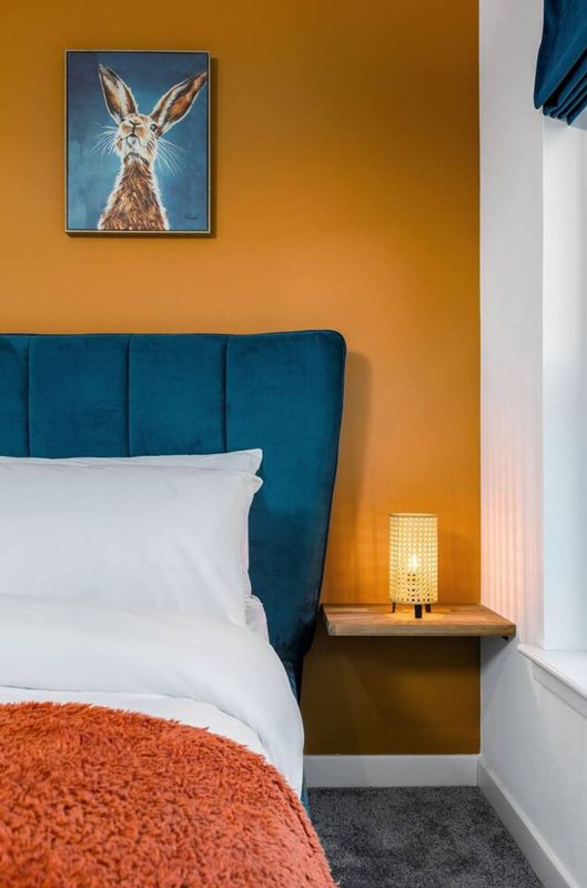 Bedroom with teal upholstered headboard, white pillows, orange throw blanket, bedside lamp and framed animal artwork