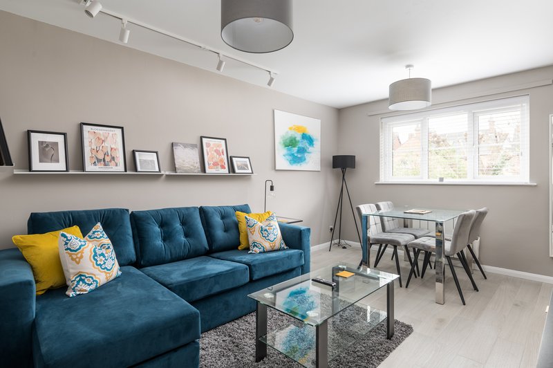 Modern living room with blue sectional sofa, yellow cushions, glass table, dining area and artwork on walls