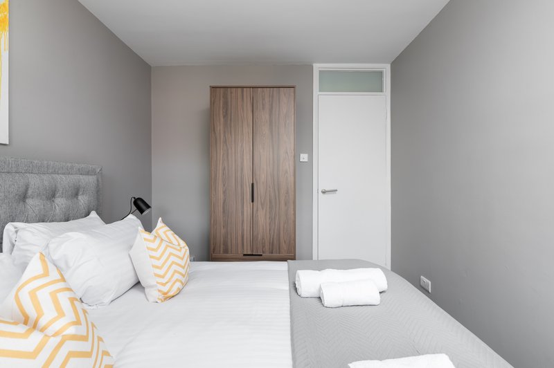 Modern bedroom with double bed, white bedding, yellow patterned cushions, wooden wardrobe and white door