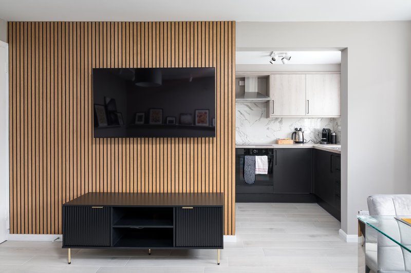 Open-plan living space with wall-mounted TV, black media console, wooden slat accent wall, and kitchen area visible