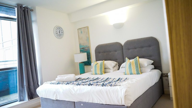 Bright bedroom with double bed, grey headboard, white bedding, decorative pillows and wall art
