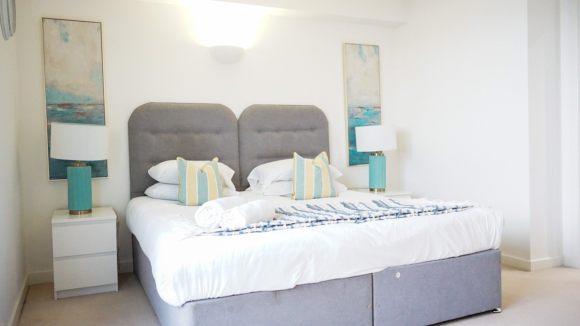 Bright bedroom with grey upholstered bed, white bedding, striped cushions and turquoise bedside tables