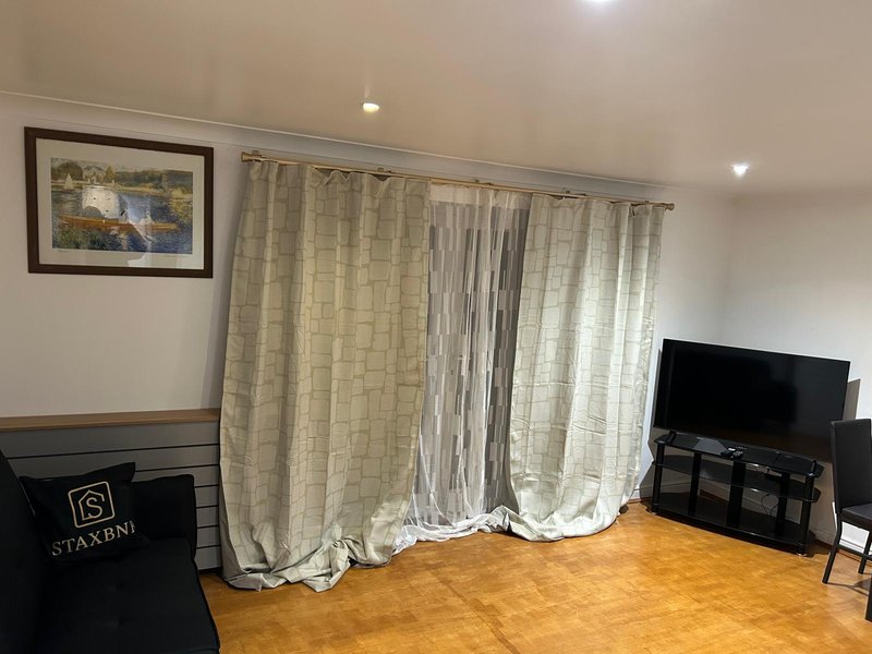 Living room with wooden floor, large windows with cream curtains, TV, and framed wall art