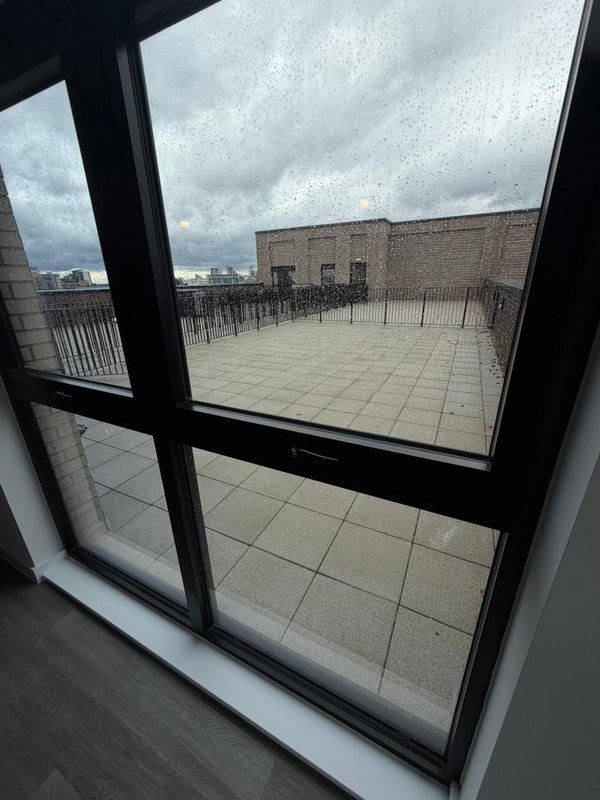 View through black-framed window to rooftop terrace with paving and railings