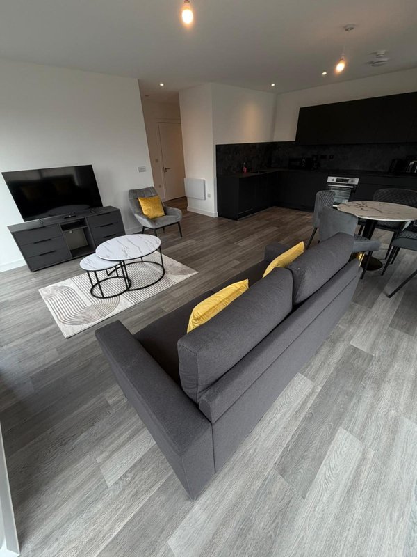 Modern open-plan living room with grey sofa, TV, kitchen area and yellow accent cushions