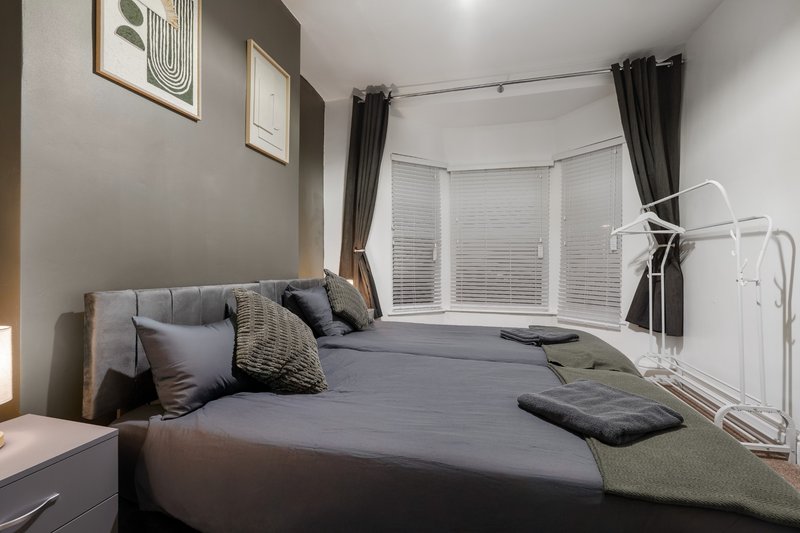 Modern bedroom with double bed, grey bedding, cushions, bay window with black curtains and white blinds