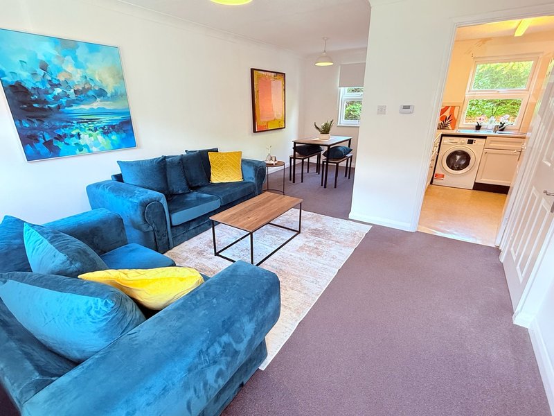 Open-plan living room with blue upholstered sofa, armchair, wooden coffee table, and kitchen visible through doorway