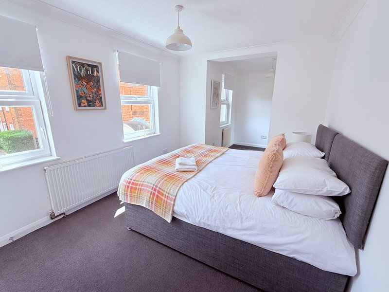Bright bedroom with double bed, grey upholstered headboard, plaid throw, and windows with radiator