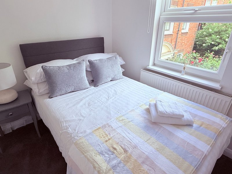 Bedroom with double bed, purple headboard, cushions, bedside table and window with garden view
