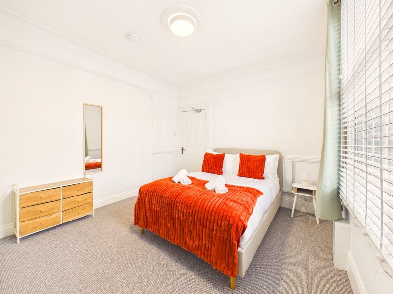 Bright bedroom with double bed, orange bedding, wooden dresser, mirror and window blinds