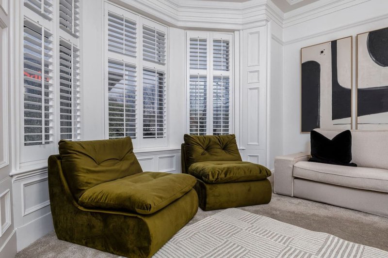 Modern living room with olive-green armchairs, white shuttered windows, cream sofa and abstract wall art