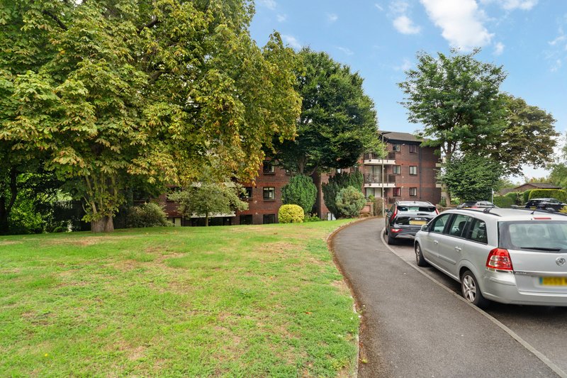 Brick apartment building with mature trees, green lawn, and driveway with parked cars