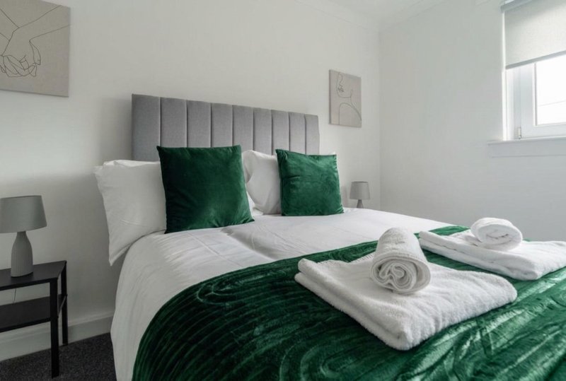 Modern bedroom with grey upholstered headboard, white and green bedding, rolled towels, and side lamp