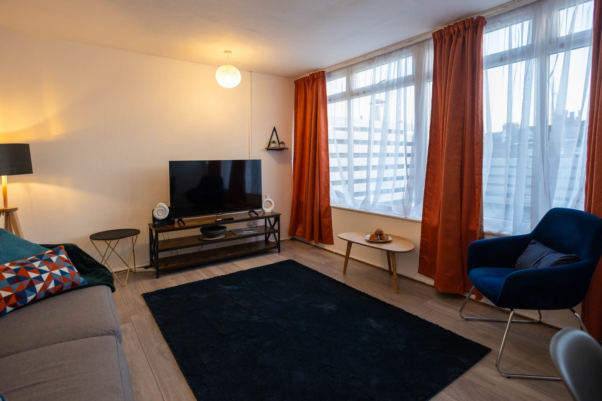 Modern living room with TV, blue armchair, gray sofa, orange curtains and city views