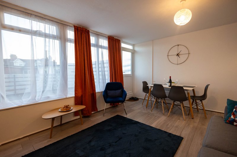 Modern living space with dining table, blue chair, large windows with red curtains, wooden flooring