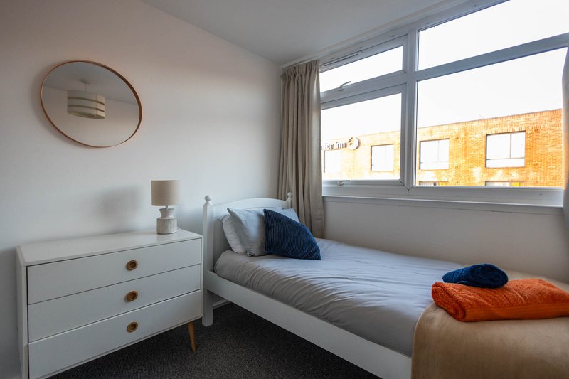 Bedroom with single bed, white dresser, round mirror, large window and orange throw