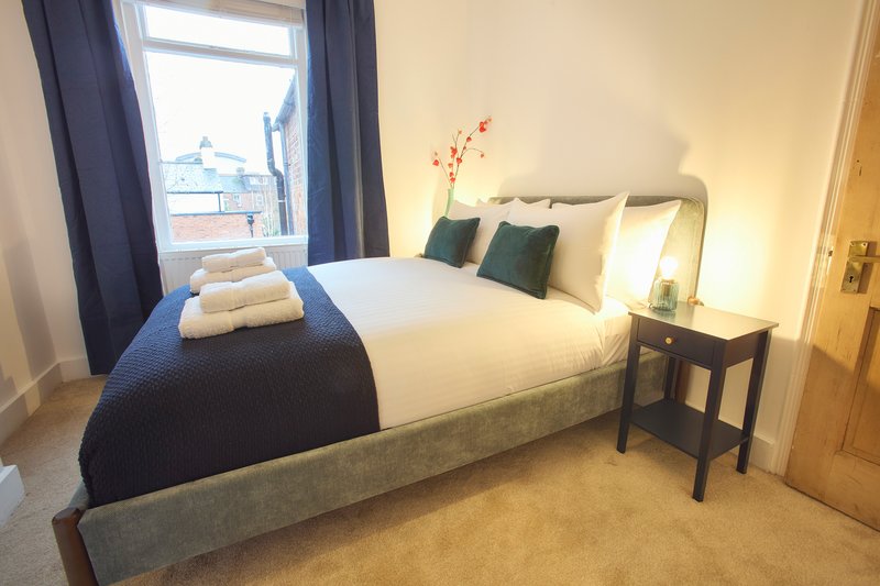 Bright bedroom with double bed, navy bedding, green cushions, bedside table and window with curtains