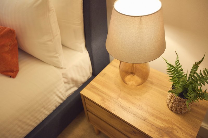 Bedside table with wooden lamp, potted plant, and bed with pillows visible