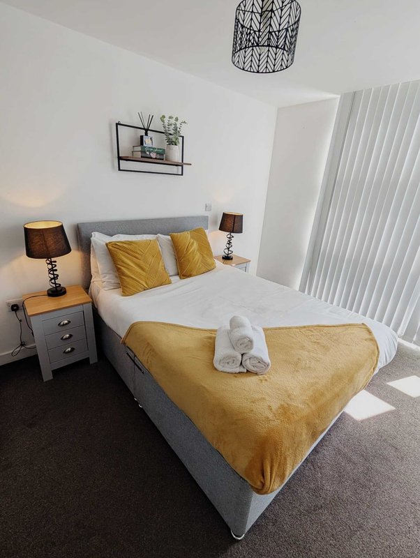 Bright bedroom with double bed, golden cushions, bedside tables, pendant light and vertical blinds