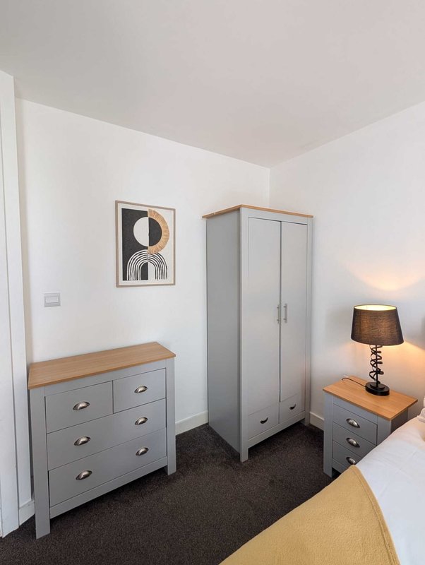 Bedroom corner with grey wardrobe, chest of drawers, bedside lamp and framed wall art