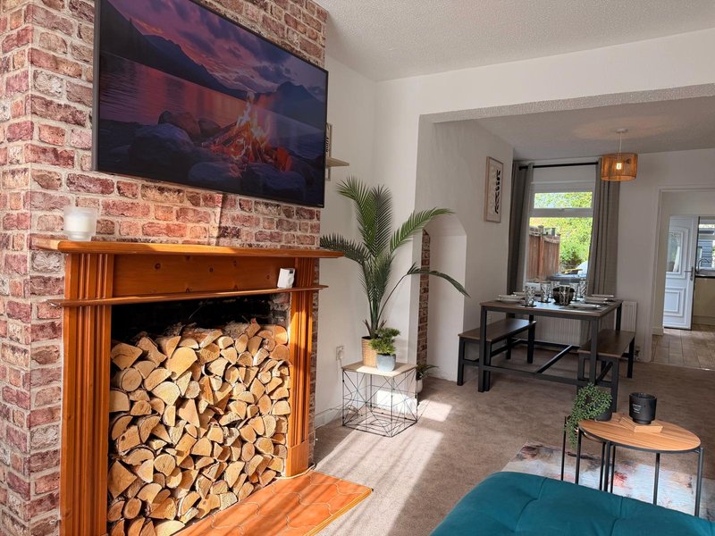 Living room with brick fireplace, wood storage, mounted TV, plants, and workspace visible through doorway