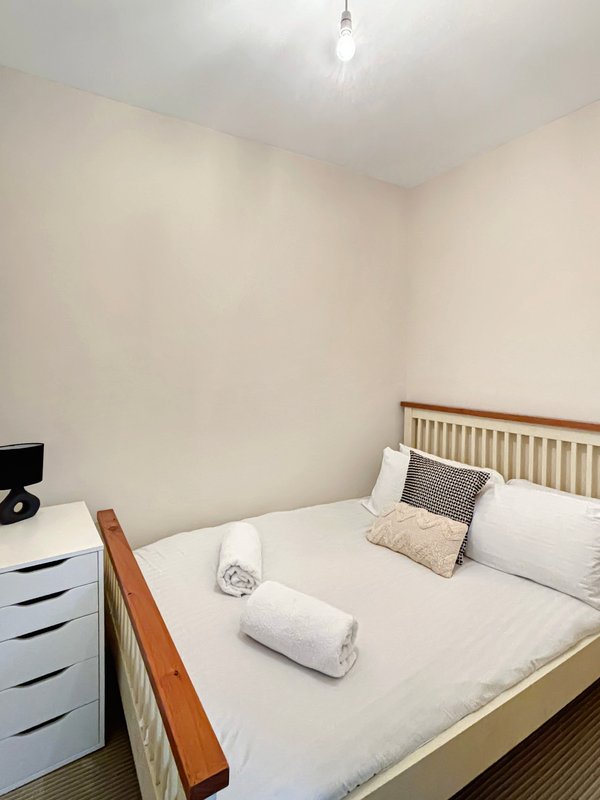 Bedroom with wooden bed frame, white bedding, rolled towels, decorative cushions and white chest of drawers