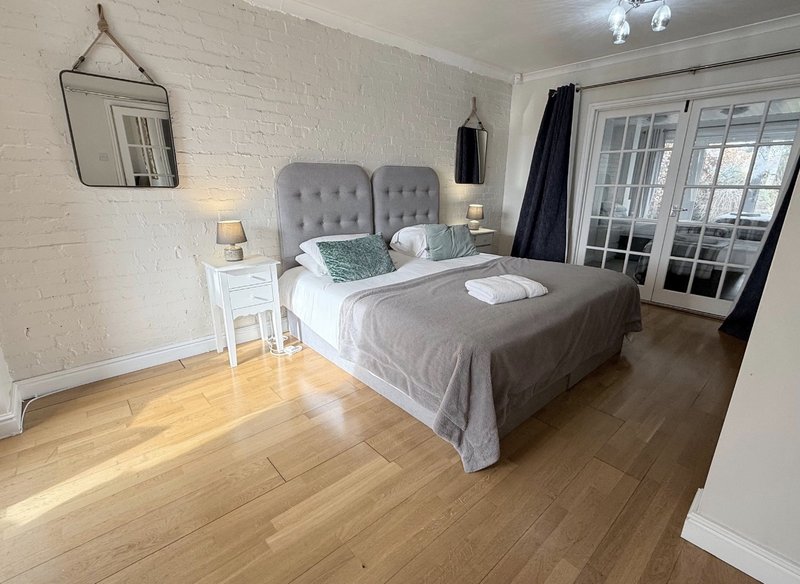 Bright bedroom with grey upholstered bed, wooden flooring, white bedside table and glass doors