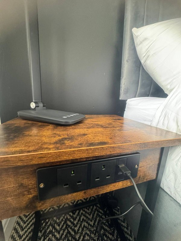 Wooden desk with power strip and router, white pillows visible on bed beside
