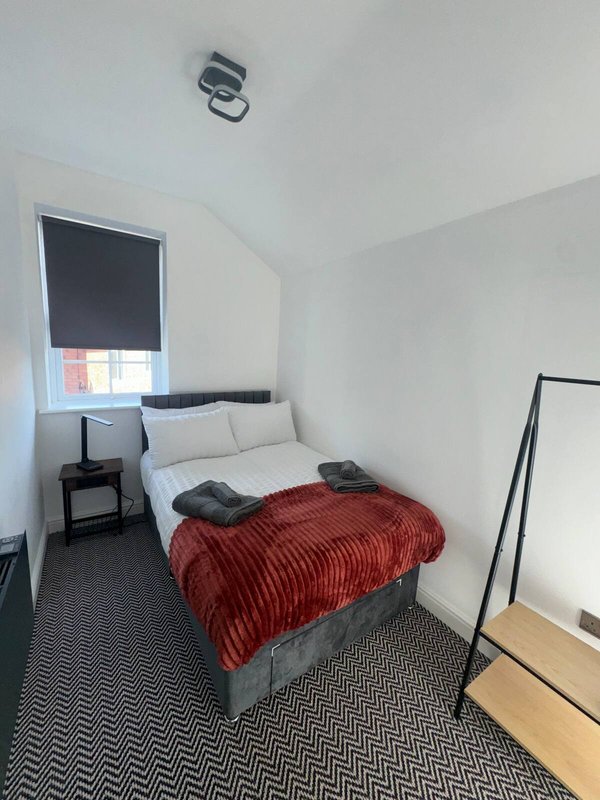 Compact bedroom with double bed, red blanket, window with blind and bedside table