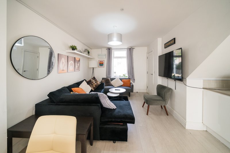 Modern living room with dark L-shaped sofa, wall-mounted TV, round mirror, and orange accent cushions