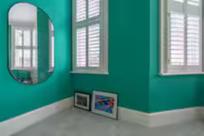 Bright teal bedroom with oval mirror, white shuttered windows and grey flooring