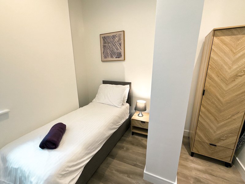 Compact bedroom with single bed, white bedding, dark headboard, wooden wardrobe and framed wall art