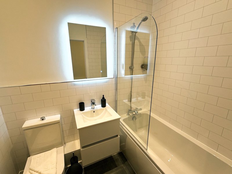 Modern bathroom with illuminated mirror, white subway tile, sink vanity, toilet, and shower bathtub