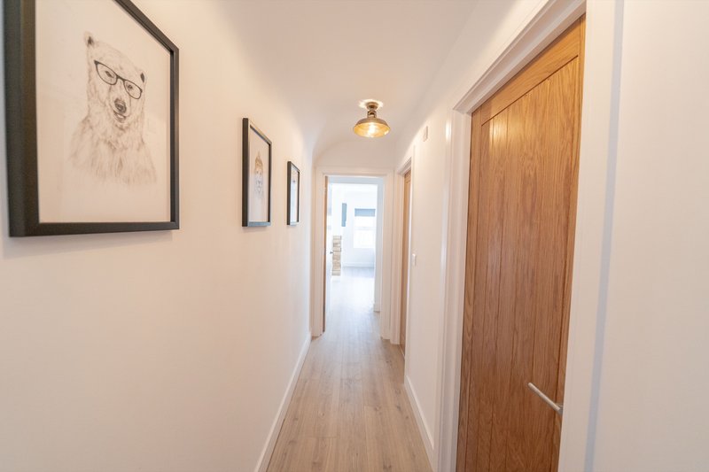Long hallway with wooden flooring, framed artwork on walls, pendant light fixture and wooden door.