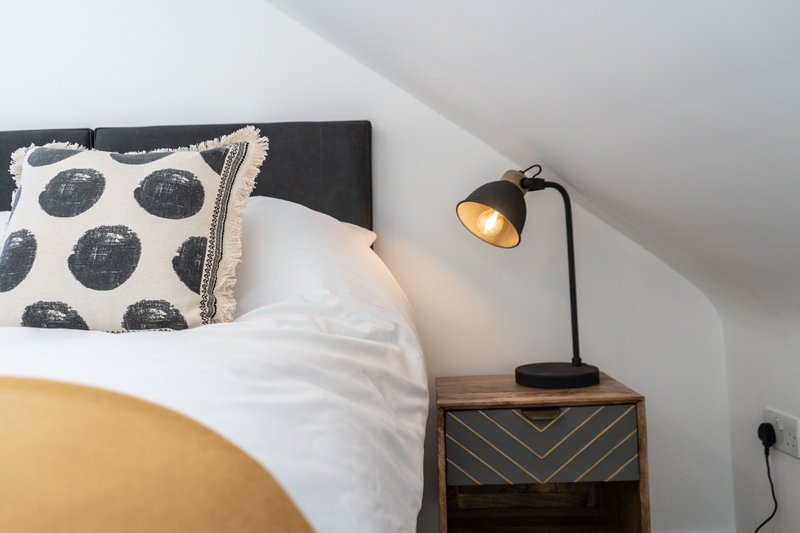 Bedroom with black headboard, patterned cushion, white bedding, wooden nightstand and black desk lamp