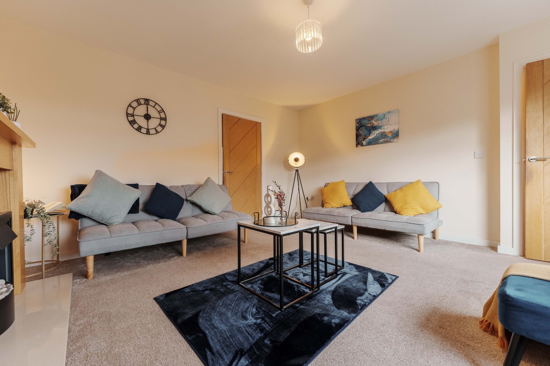Open-plan living area with two grey sofas, yellow and blue cushions, coffee table, wall clock and pendant lighting