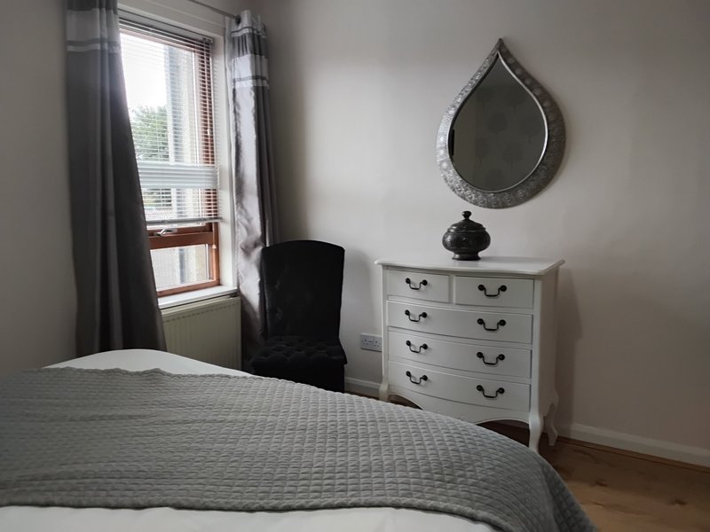 Bedroom with double bed, white dresser, black chair, teardrop mirror and window with curtains