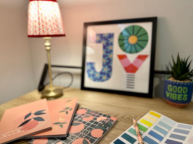Wooden desk with lamp, framed art, notebooks, color swatches, and potted plant