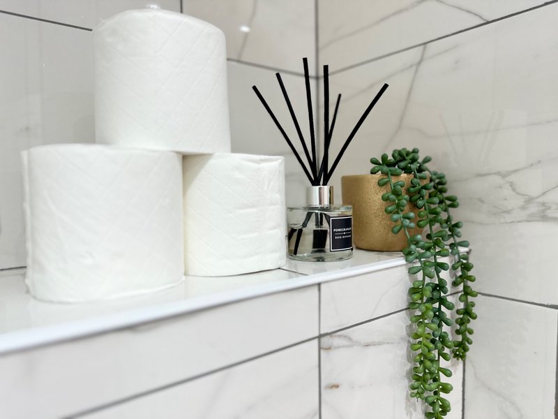 White bathroom shelf with toilet paper rolls, reed diffuser, potted plant and marble tile backsplash