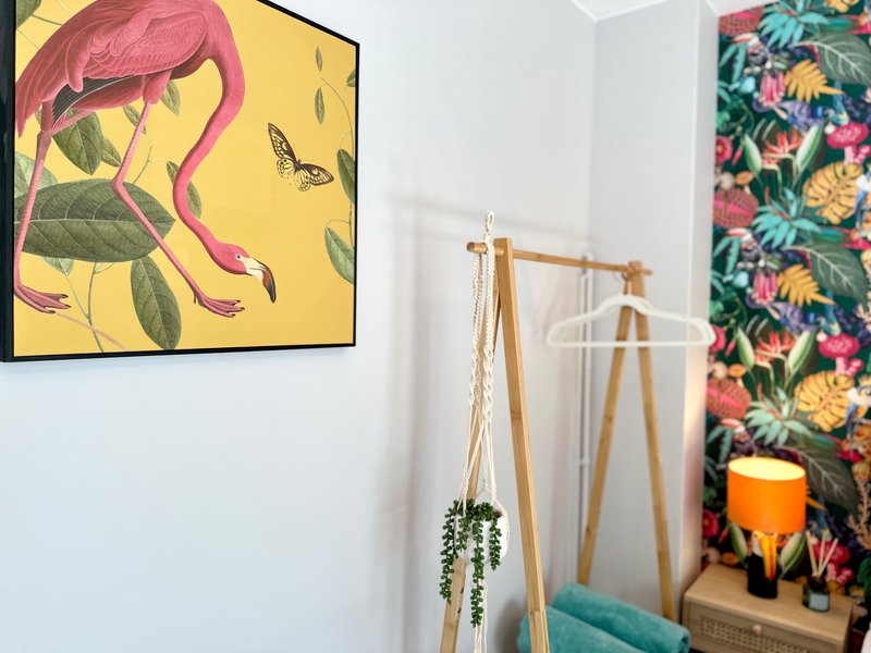 Modern bedroom corner with framed botanical artwork, wooden coat stand, hanging plant, and floral wallpaper