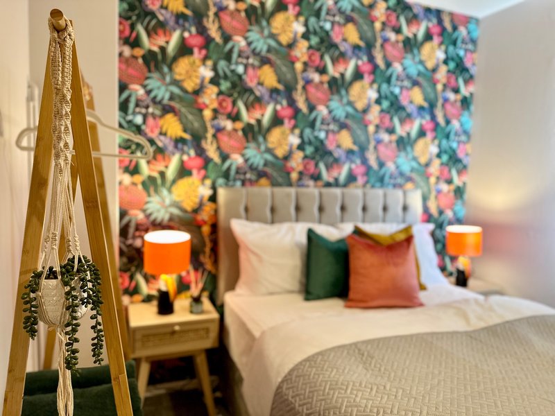 Bedroom with colorful floral wallpaper, double bed with green and orange pillows, wooden tripod with hanging plant, and bedside lamps