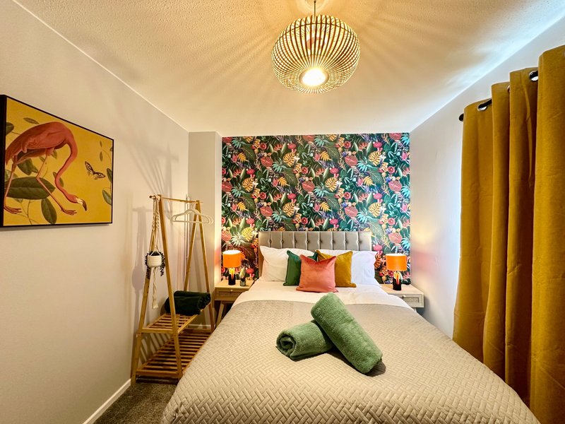 Bright bedroom with double bed, floral headboard wallpaper, colorful cushions, and gold ceiling light fixture