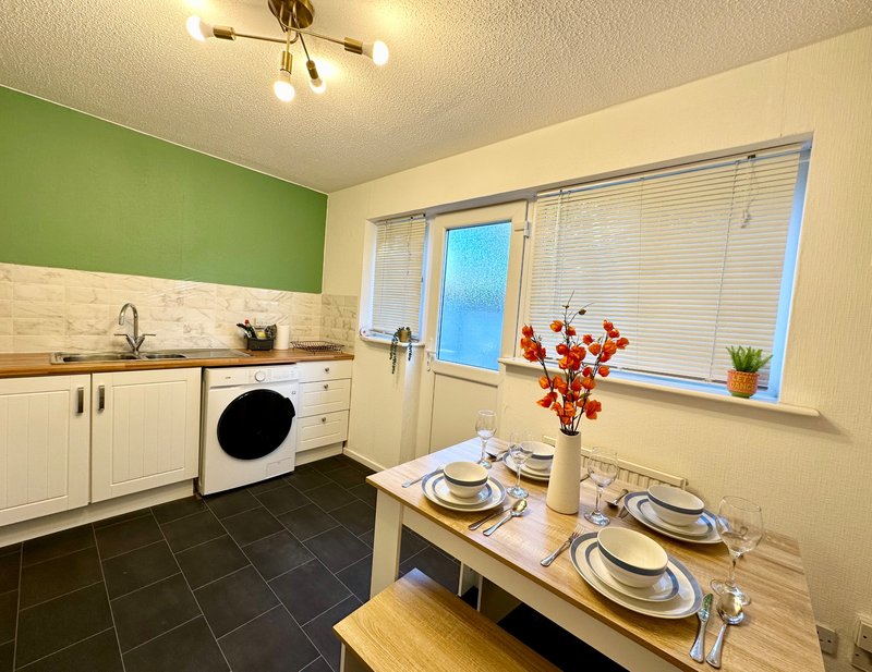 Kitchen diner with cream cabinets, green accent wall, dining table set for two, washing machine and windows