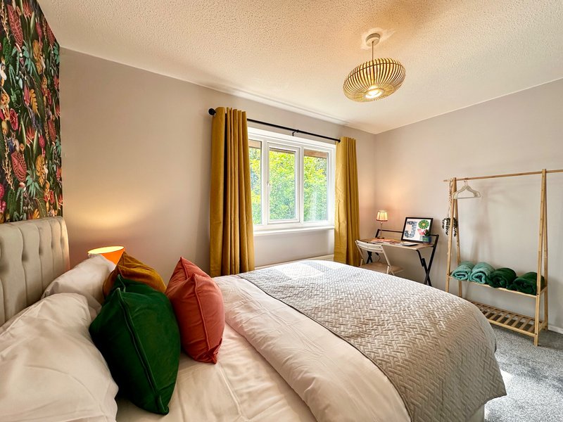 Bright bedroom with double bed, colorful cushions, desk, window with gold curtains and pendant light