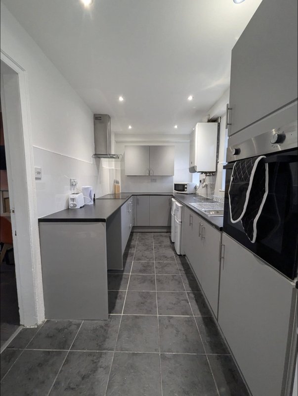 Modern galley kitchen with white cabinets, black countertop, stainless steel appliances and dark tile flooring.