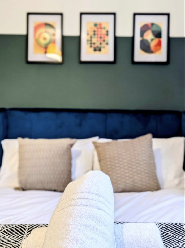 Bedroom with blue upholstered headboard, white bedding, pillows, and three framed abstract artworks on green wall