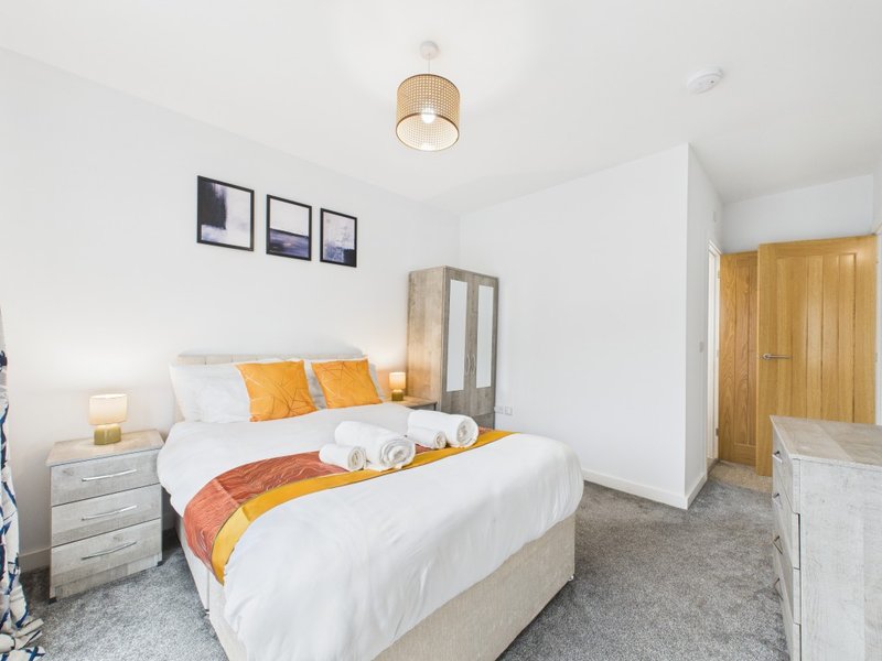 Modern bedroom with double bed, white bedding, orange and yellow accents, wooden wardrobe and framed wall art