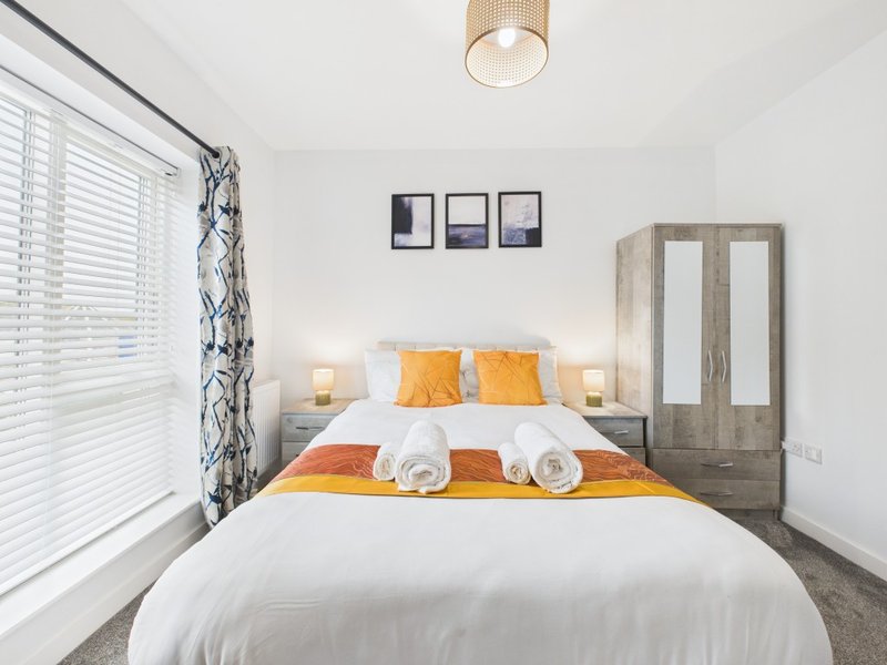 Bright modern bedroom with double bed, yellow and orange bedding, wooden wardrobe, and window blinds